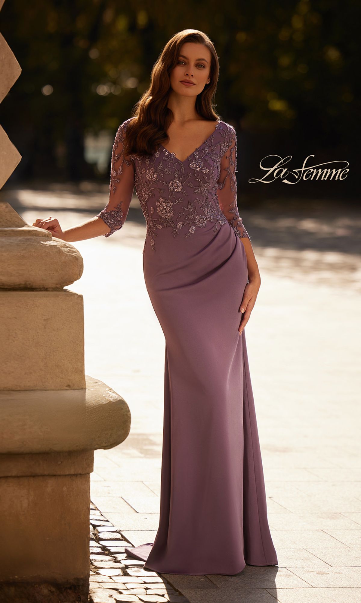 A woman in a dusty purple La Femme 32738 long sleeve beaded formal gown stands outdoors by a stone structure. The text